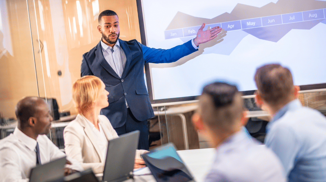Photo of a man making a presentation pointing to a screen with a graphic on it.